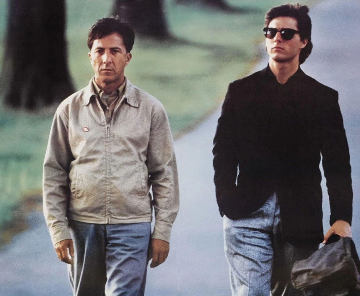 Two men walking side by side, one in a light jacket and the other wearing sunglasses and carrying a bag.